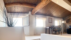 A spacious loft with exposed wooden beams, a staircase, a dining area with chairs, and a stone wall.