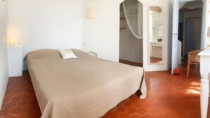A bedroom with a bed, a nightstand, a wall-mounted lamp, a door leading to another room, and a tiled floor.