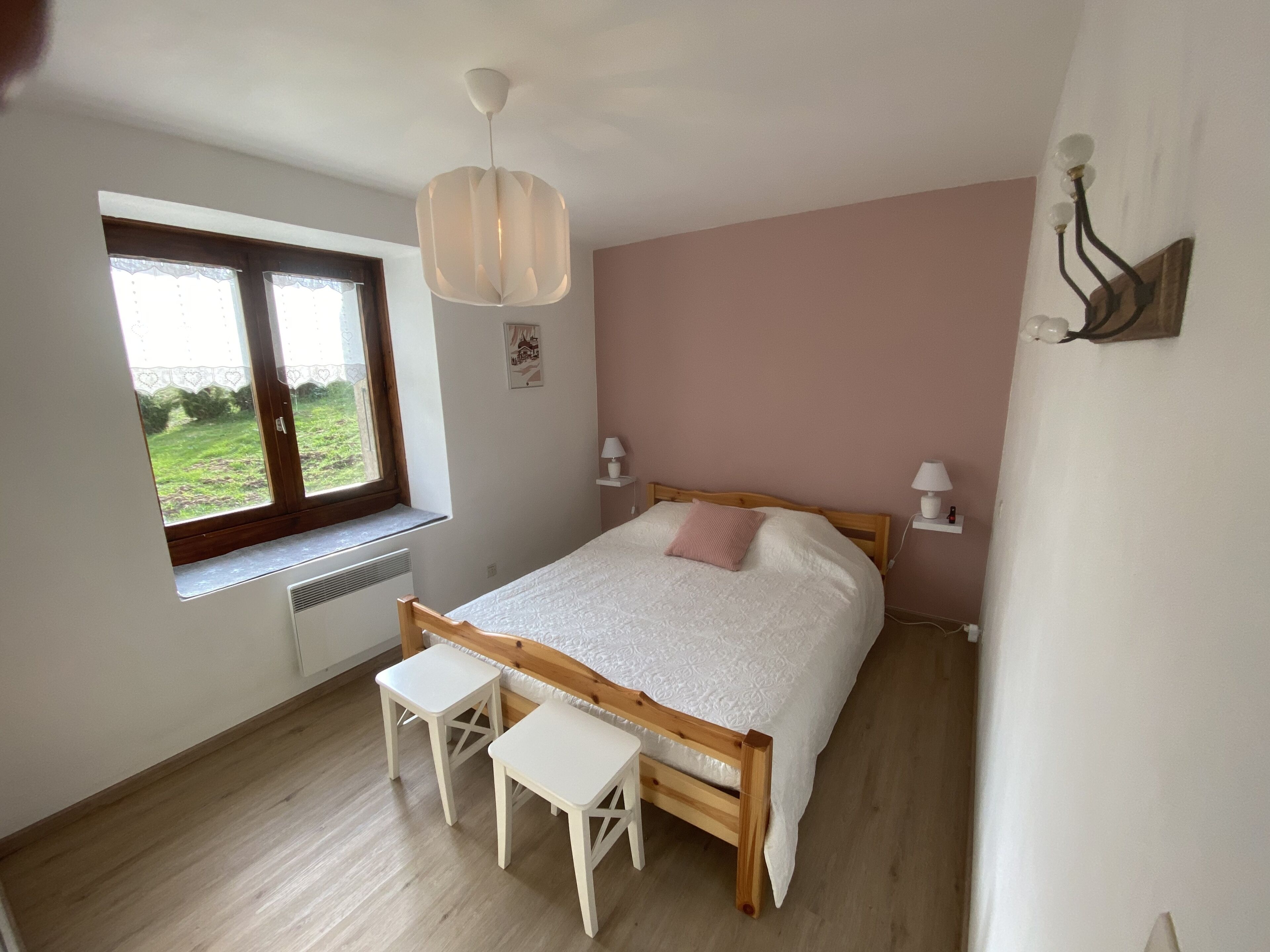 Une chambre avec un lit en bois, deux tables de chevet blanches, une fenêtre donnant sur un paysage verdoyant, un luminaire au plafond et une lampe fixée au mur.