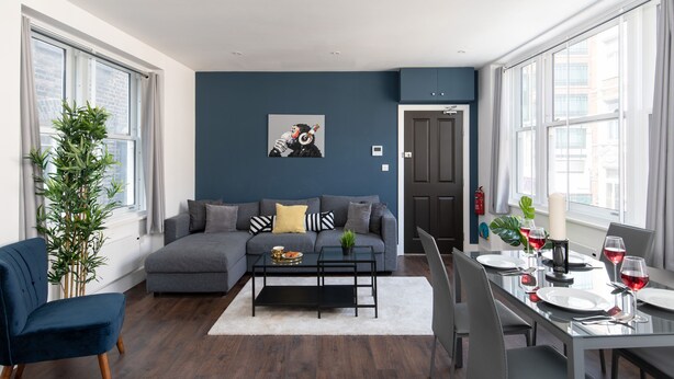 A modern living room with a grey sofa, a coffee table, and a dining area with a glass table set for a meal. - home.ly London Liverpool Str Apartments (London)