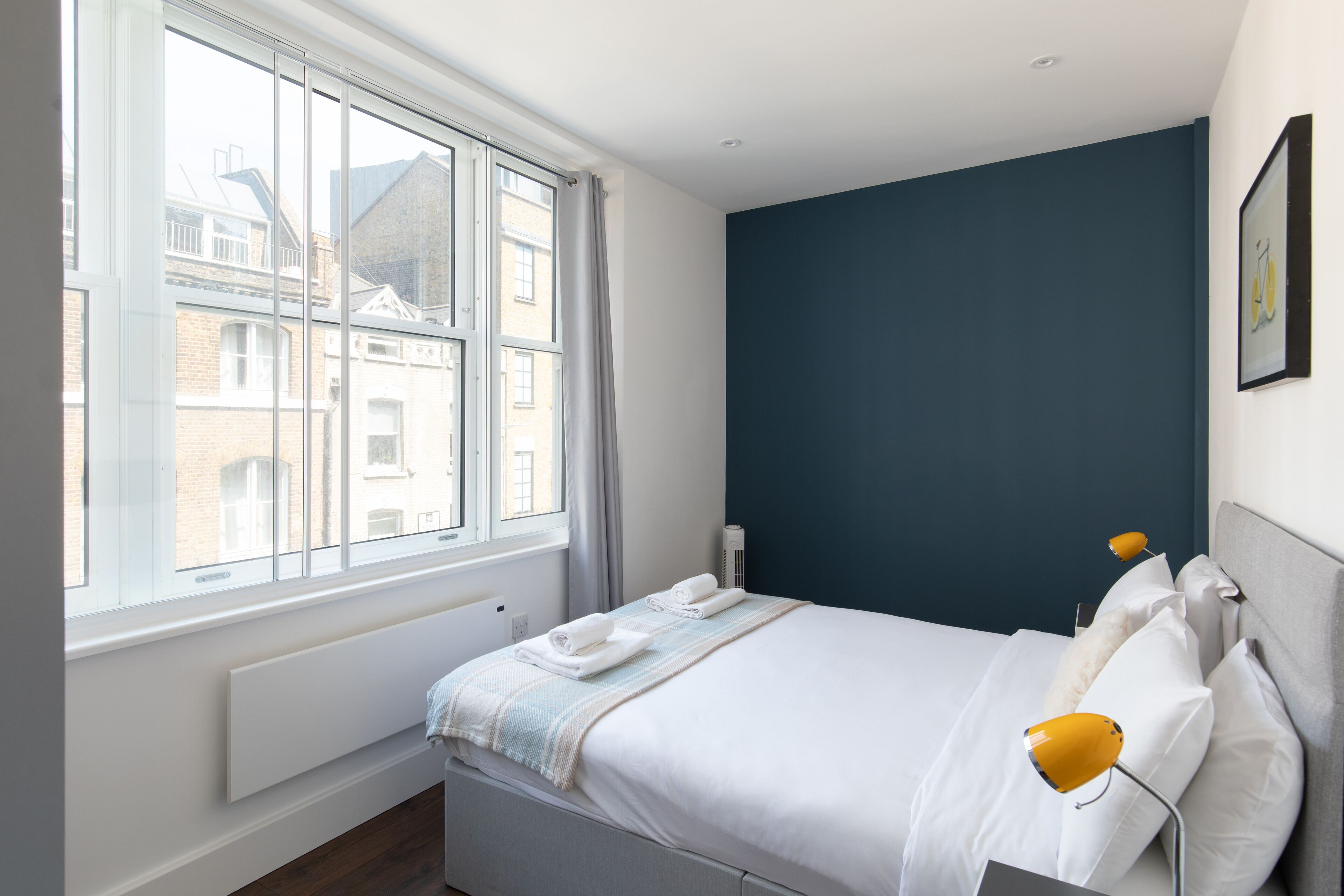 A bedroom with a large window, a bed with white linens, a blue accent wall, and a yellow bedside lamp.