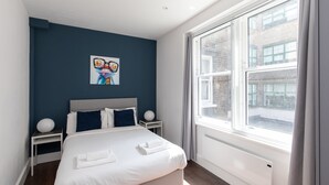 A modern hotel room with a large bed, a nightstand, a wall art piece, and a large window with a view of the city. - home.ly London Liverpool Str Apartments (London)