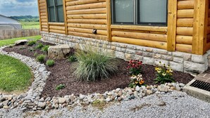 A log cabin with two windows, a stone foundation, and a landscaped garden with rocks and plants.