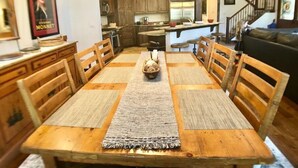 Un comedor con mesa y sillas de madera, una alfombra en el centro y una cocina al fondo.