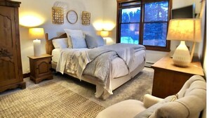 Un dormitorio con cama, lámparas de noche, mesita de noche, cómoda y ventana con vista a árboles.