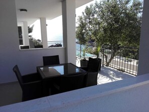 Een balkon met een glazen tafel en stoelen, uitzicht op een zwembad en bomen, en een witte reling.