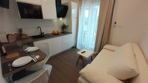 Een moderne keuken met een ingebouwde magnetron, een spoelbak en een fruitschaal. Er is een zithoek met een tafel gedekt voor twee personen, een witte bank en een klein bijzettafeltje.