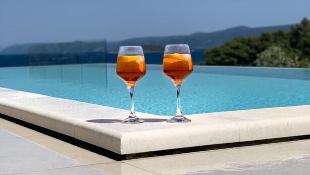 Twee glaasjes amberkleurige vloeistof bij een zwembad, met een helderblauwe lucht en verre bergen.