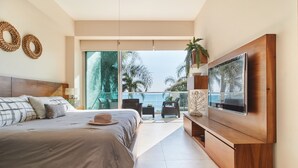 A modern bedroom with a large bed, a TV, and a balcony with a view of palm trees and the ocean.