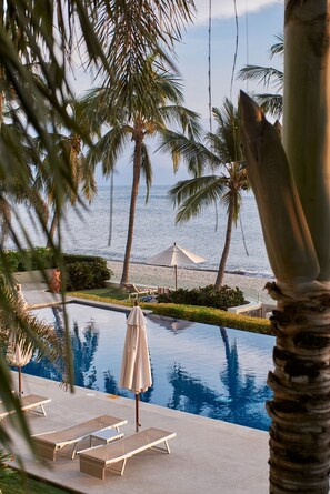 A pool area with lounge chairs and umbrellas, palm trees, and a view of the ocean.