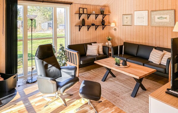 A modern living room with a sofa, armchair, and coffee table, wooden floors, and a large window with a view of a garden.