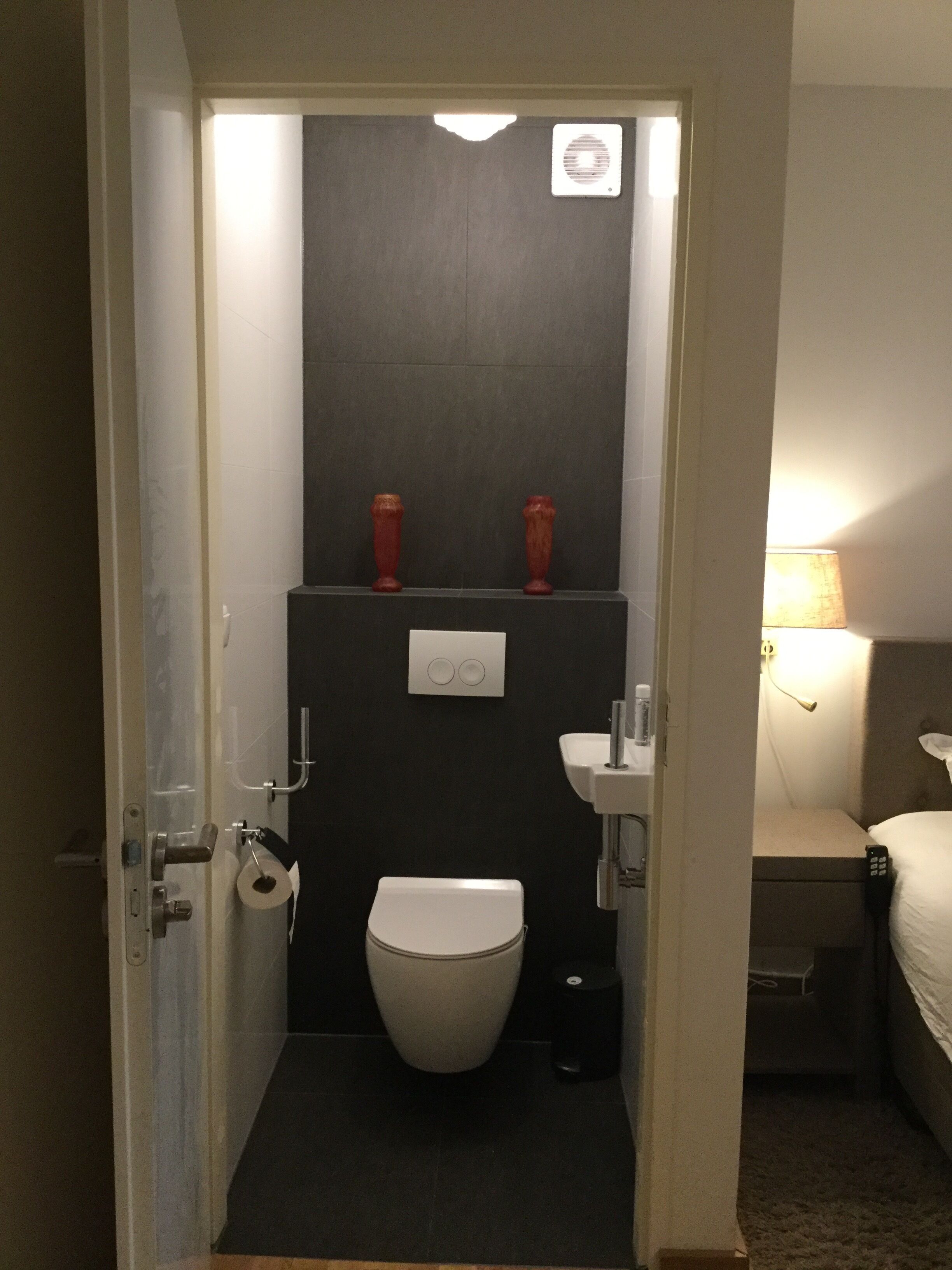 A compact bathroom with a toilet, a sink, and a mirror. There is a towel dispenser and a small trash bin.