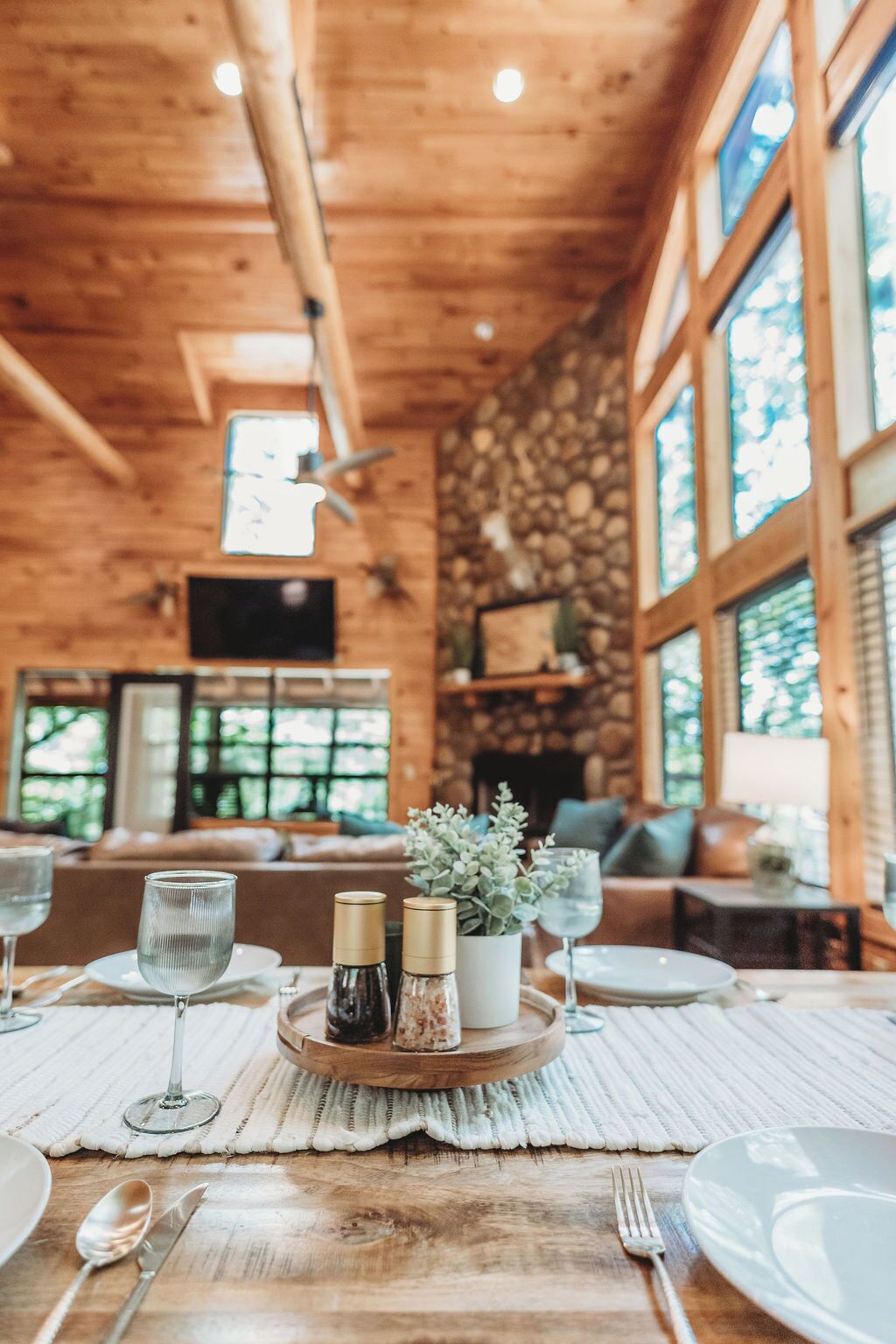 A wooden interior with a dining table set for two, featuring a central wooden tray with condiments and a potted plant.