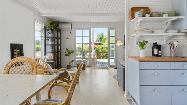 Une cuisine lumineuse avec des meubles blancs, un coin repas doté de chaises en osier et une vue sur l’extérieur grâce à de grandes fenêtres.