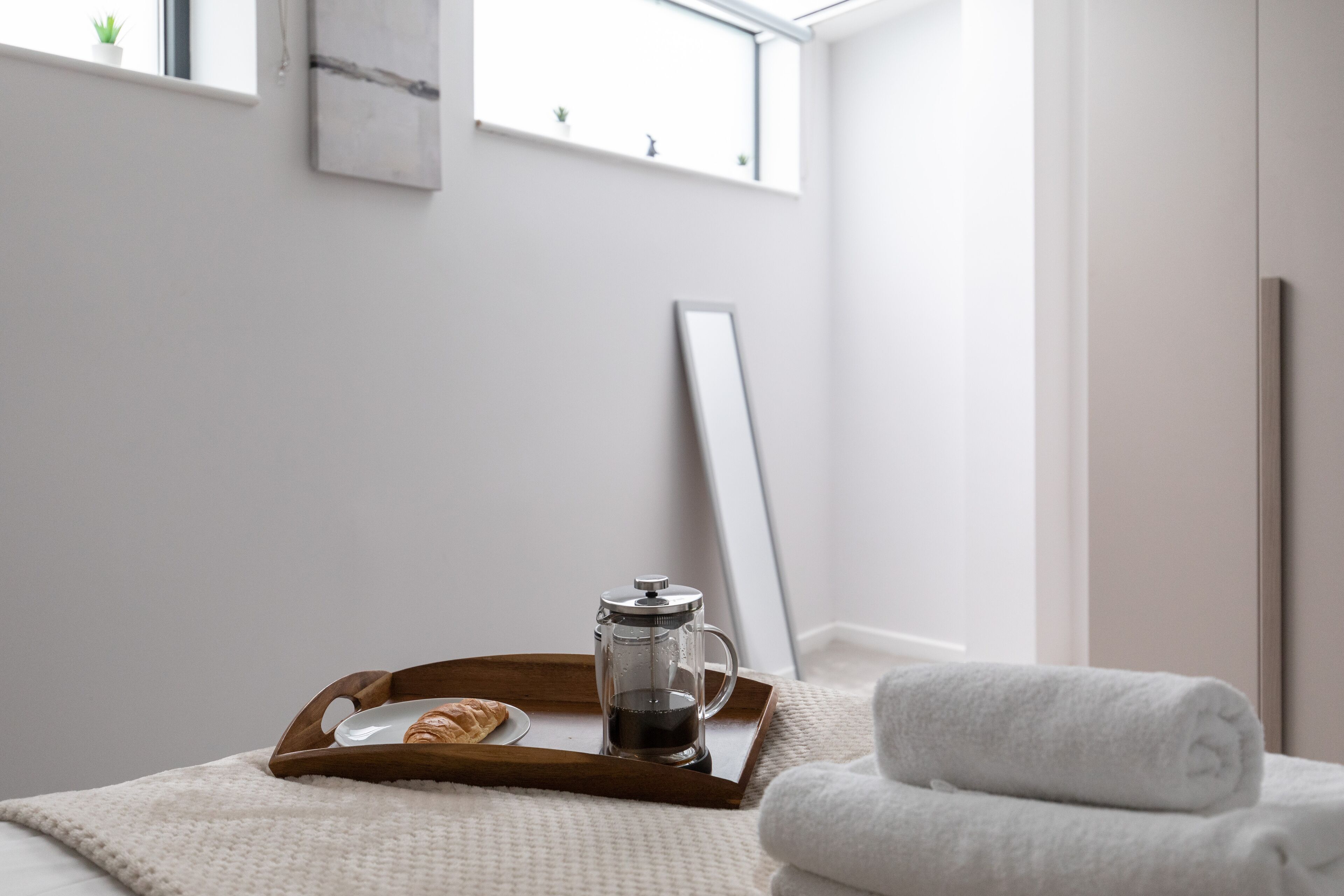 A neatly made bed with a white towel, a tray with a coffee pot and croissant, and a window with a view of the sky.