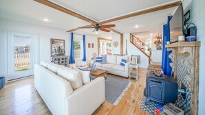 A spacious living room with a white sofa, wooden beams, a ceiling fan, and a wood-burning stove.