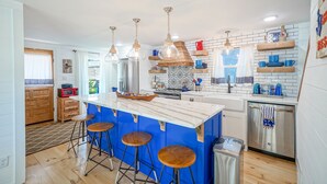 A kitchen with a central island, bar stools, hanging pendant lights, and a stainless steel dishwasher.
