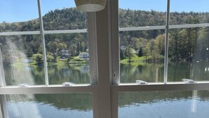A room with a large window overlooking a lake and forested hills. - Lake Front Hotel (Cooperstown)