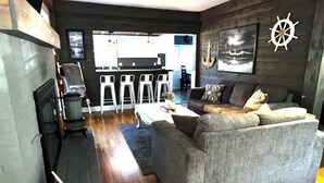 Una sala de estar con un sofá gris, piso de madera, un televisor de pantalla plana y una zona de bar al fondo.
