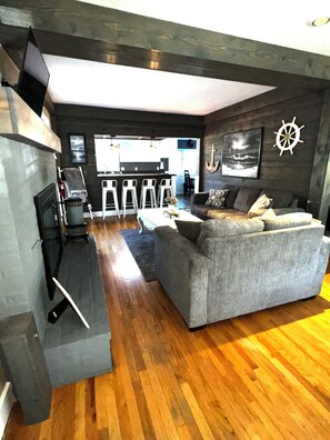 Una sala de estar con un sofá gris, piso de madera, un televisor de pantalla plana y una zona de bar al fondo.