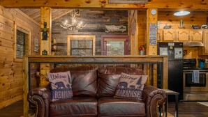 A cozy cabin interior with a leather sofa, wooden walls, a fireplace, and a kitchen area.