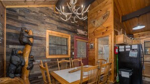 A wooden interior with a dining table, chairs, a chandelier, and a refrigerator.