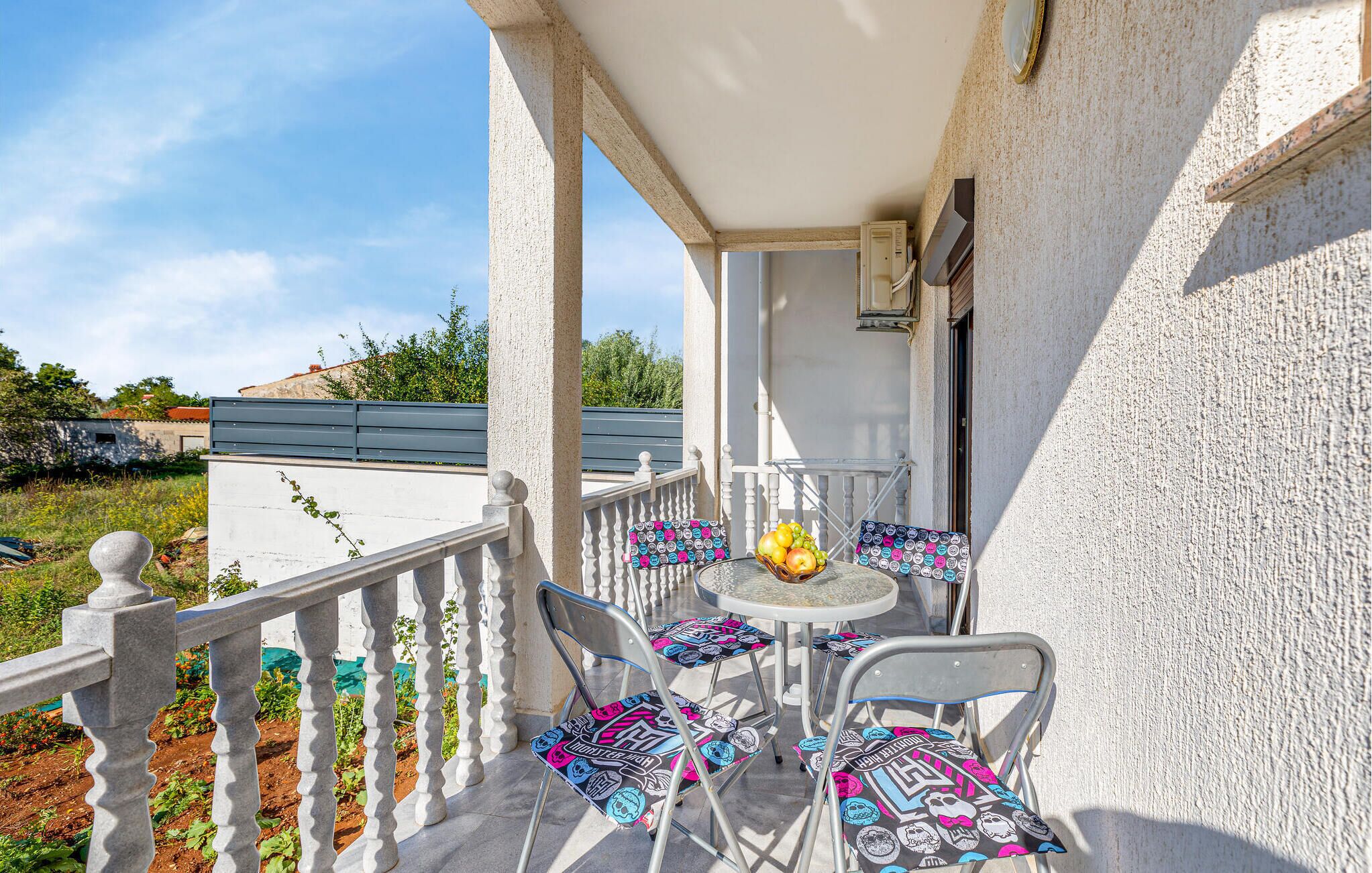 Un balcon avec une table et des chaises, un saladier rempli de fruits et une vue sur la verdure et les bâtiments.