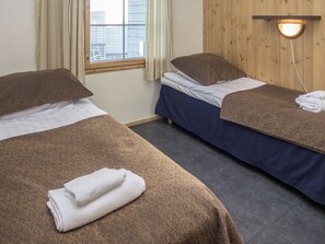 A hotel room with two beds, each with a brown headboard and a patterned bedspread. There is a window with curtains, a wall-mounted light fixture, and a wooden panel wall.