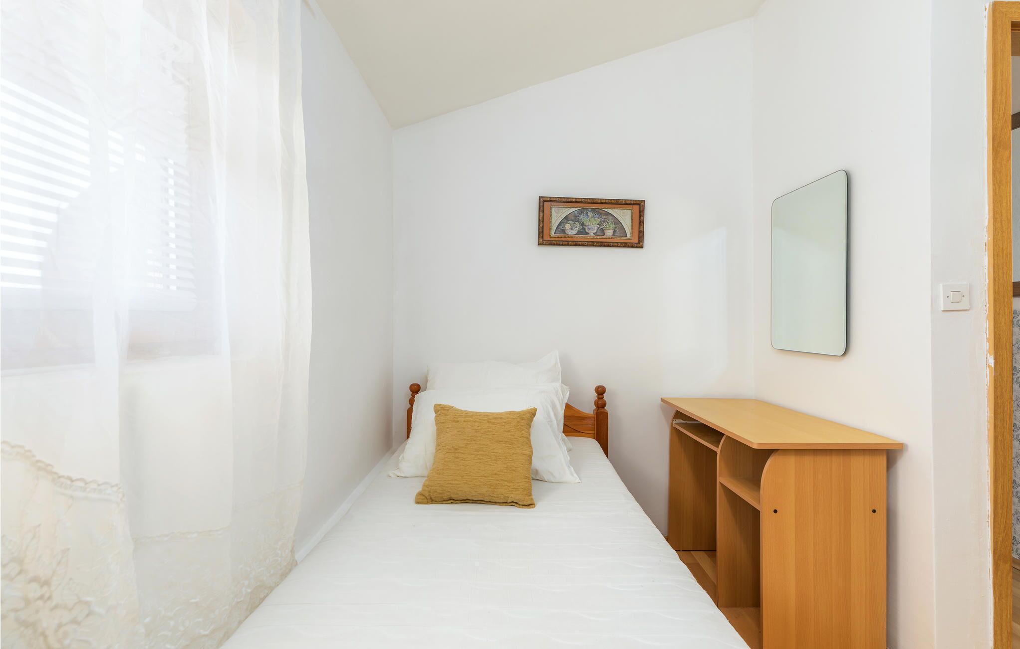 Une petite chambre à coucher, bien rangée, avec un lit simple, une table de chevet, un miroir et un tableau encadré accroché au mur.