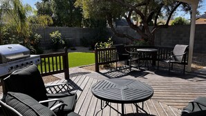 Une terrasse en bois avec des meubles d’extérieur, un barbecue et un espace jardin avec une pelouse et des arbres.