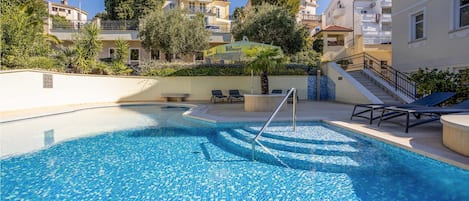 Ein Swimmingpool mit gefliestem Sonnendeck, Liegestühlen und einer Treppe, die zu einer oberen Ebene führt.