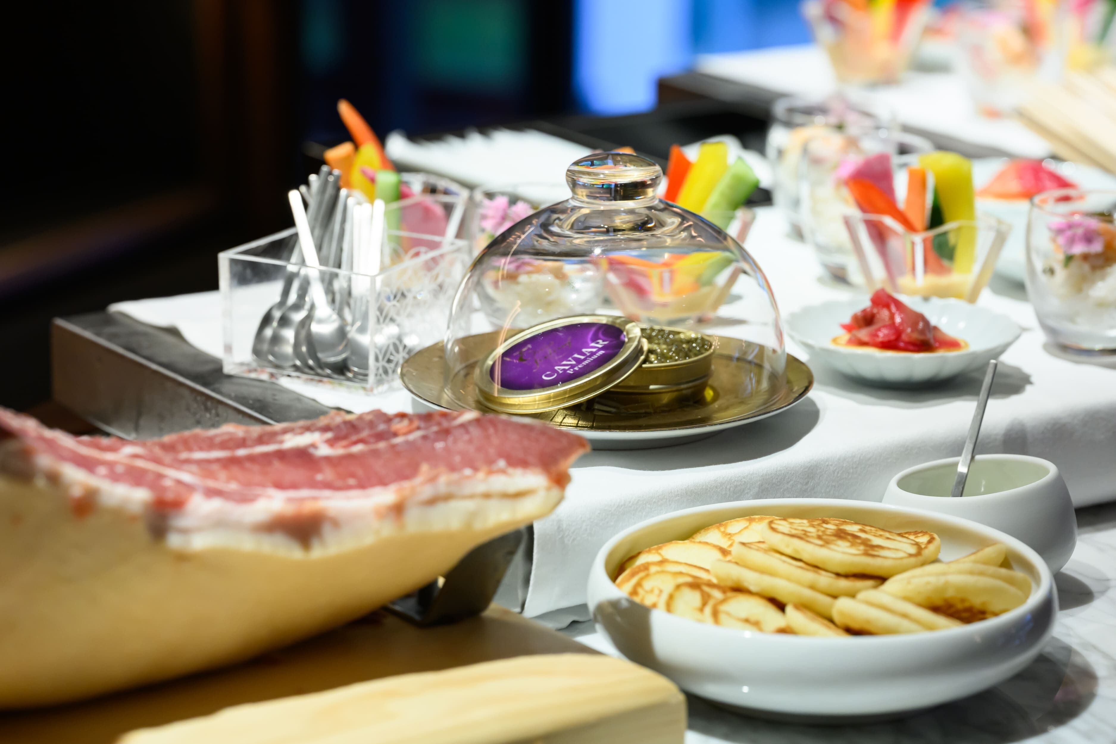 A buffet with various dishes, including a large piece of meat, a bowl of pancakes, and a glass container with a purple label.
