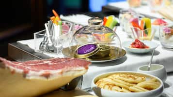 A buffet with various dishes, including a large piece of meat, a bowl of pancakes, and a glass container with a purple label.