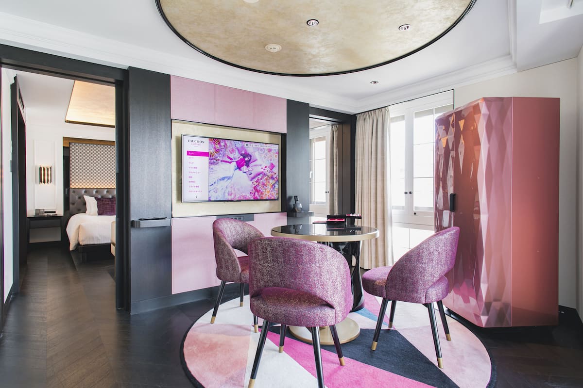 A modern hotel room with a round dining table, two purple chairs, a large abstract painting, and a bed in the background.