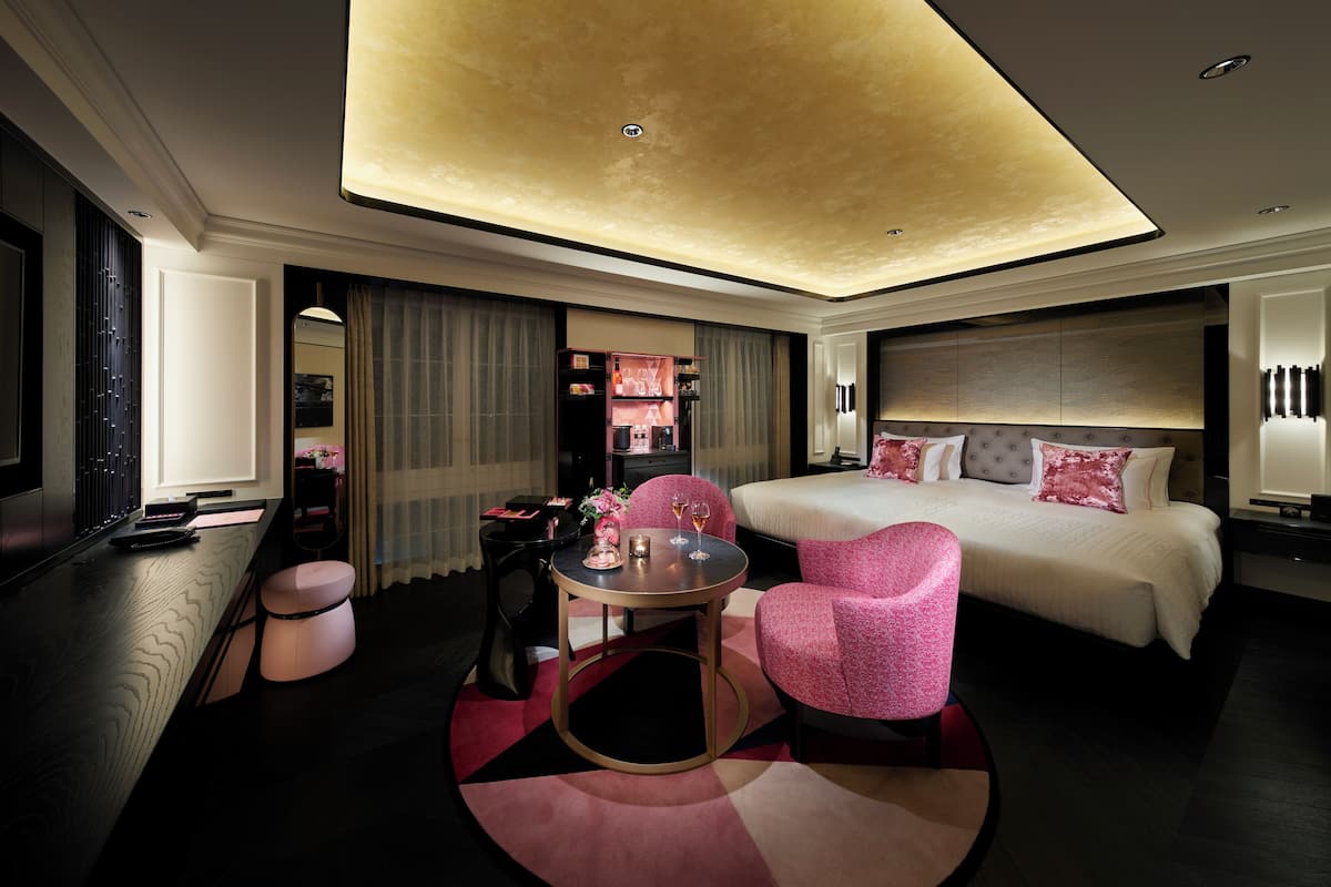 A modern hotel room with a large bed, a sitting area with a pink chair, a small round table, and a ceiling light fixture.