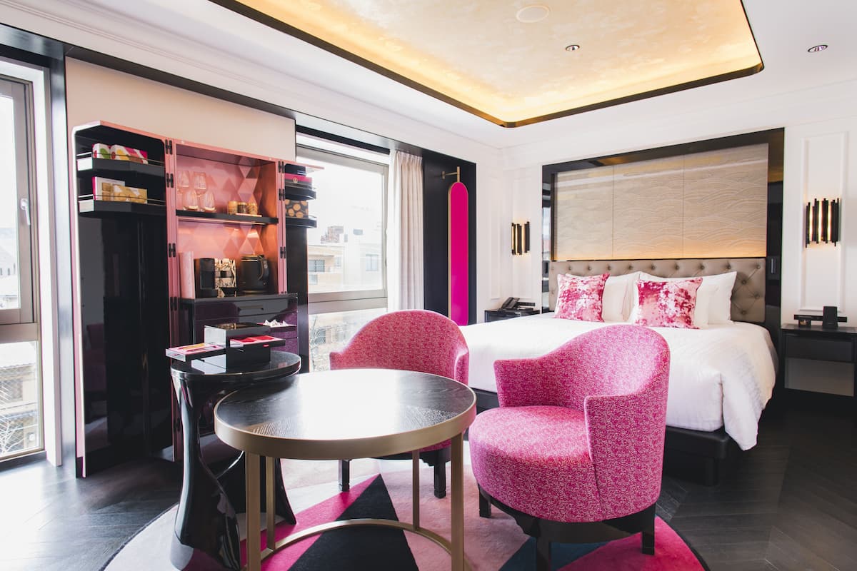 A modern hotel room with a large bed, a desk, and two pink chairs.