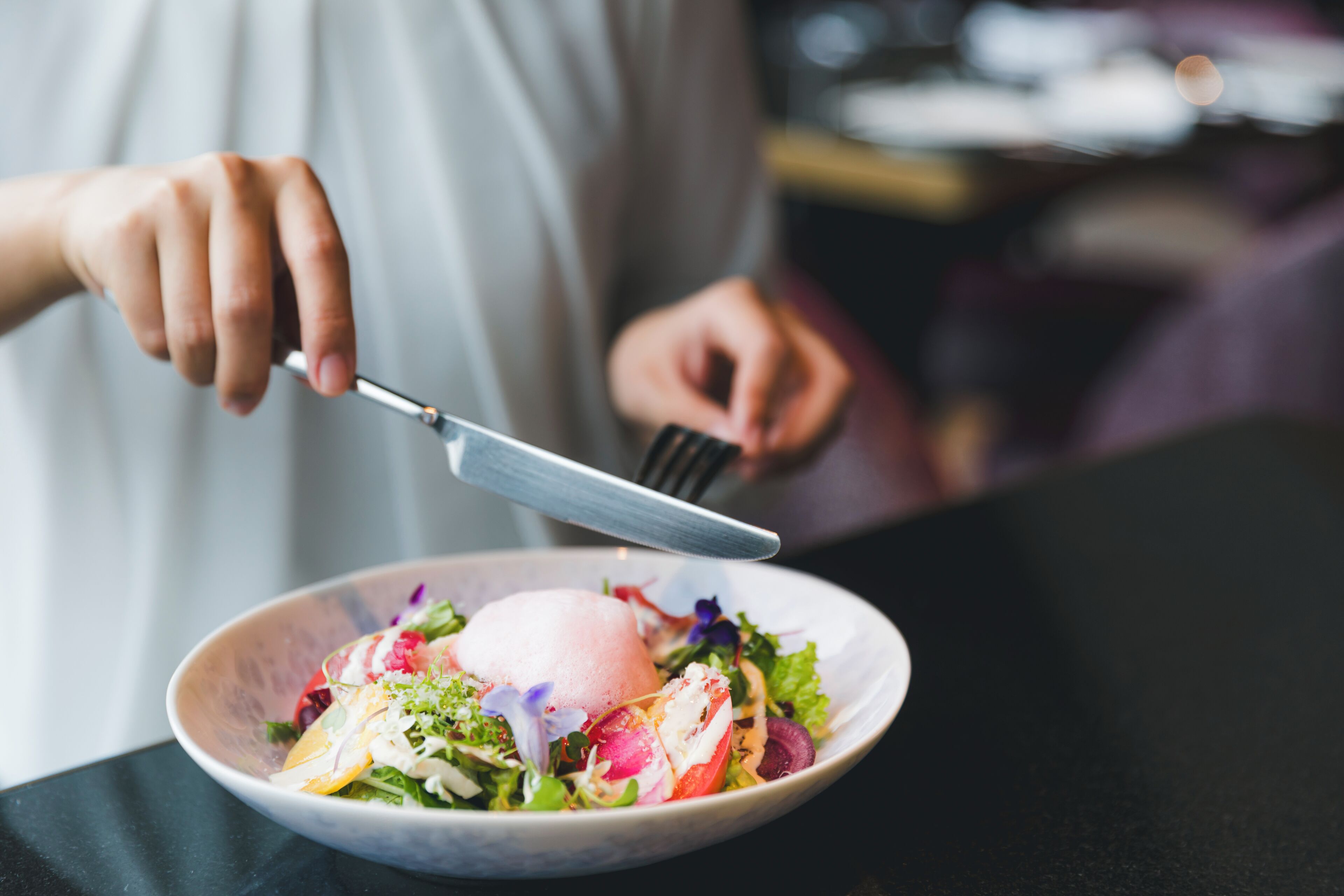 A person holding a fork and knife over a bowl of salad with a scoop of ice cream.