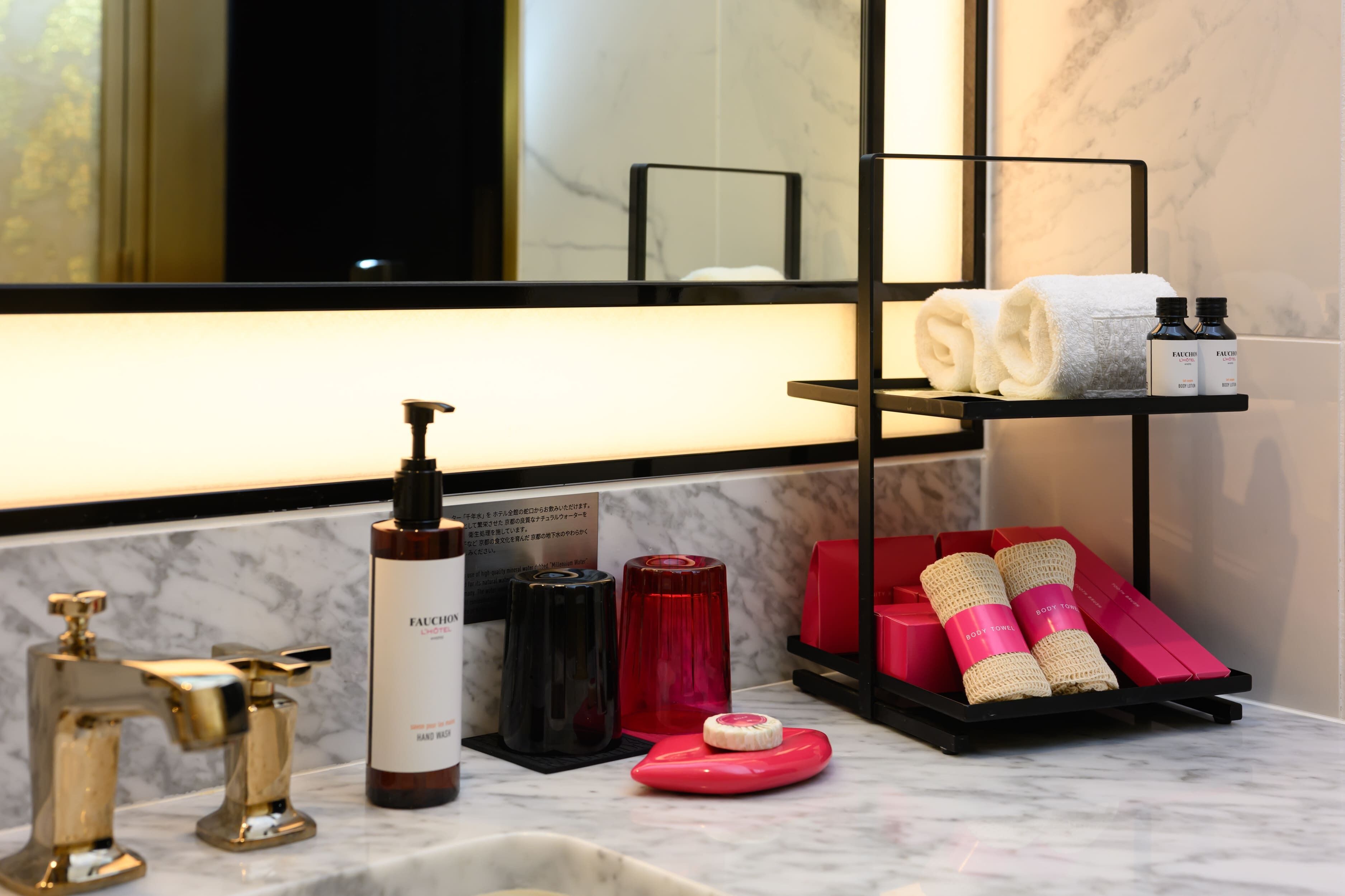 A bathroom with a marble countertop, a black towel rack, and various toiletries including a body lotion bottle, a red soap dispenser, and pink body towels.