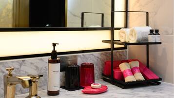 A bathroom with a marble countertop, a black towel rack, and various toiletries including a body lotion bottle, a red soap dispenser, and pink body towels.