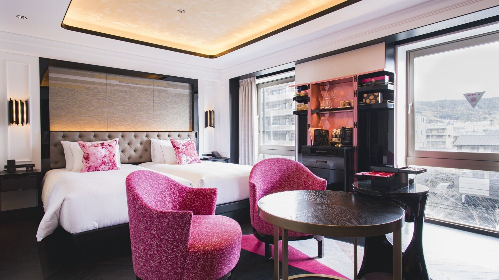 Una habitación de hotel moderna con una cama grande, dos sillas rosas, una mesita redonda y un minibar.