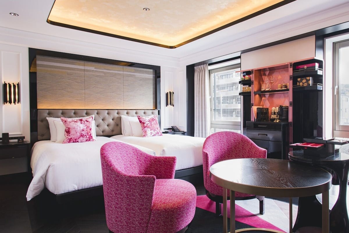A modern hotel room with a large bed, two pink chairs, a small round table, and a minibar.