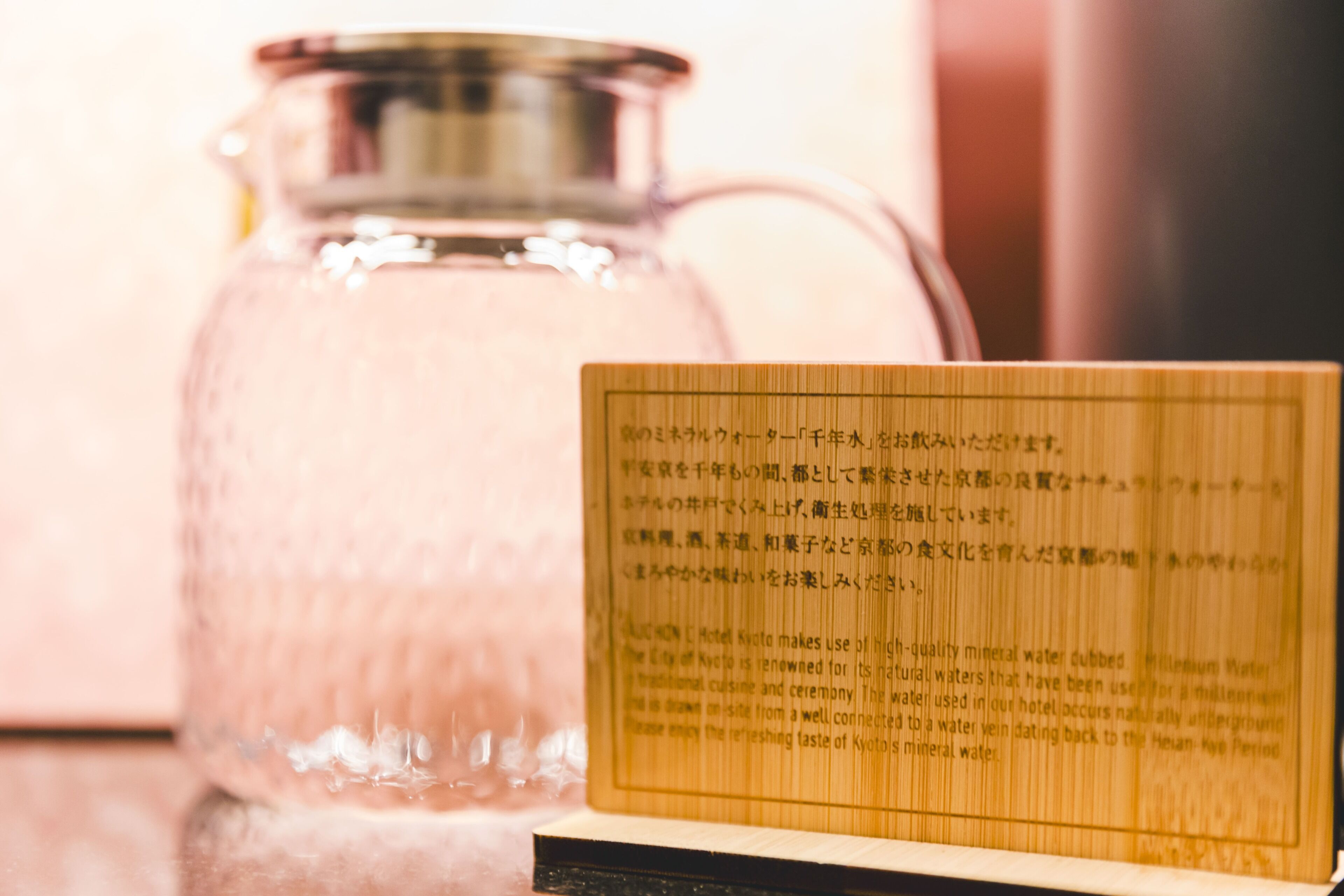 A clear glass pitcher with a metal lid, placed on a reflective surface. In front of the pitcher, there is a wooden plaque with Japanese text.
