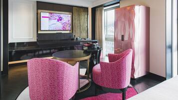 A modern hotel room with a round dining table, two pink upholstered chairs, a flat-screen TV mounted on the wall, and a large window with a view of the city.