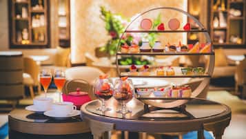 A tiered dessert display with various pastries and a tea set on a table in a warmly lit room with seating and a decorative backdrop.