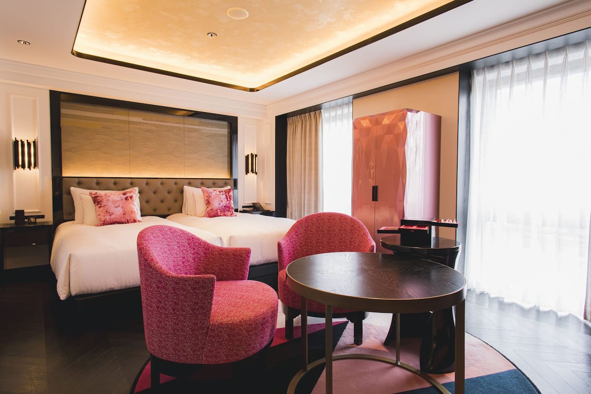 A hotel room with a large bed, two red upholstered chairs, a round wooden table, and a large window with curtains.
