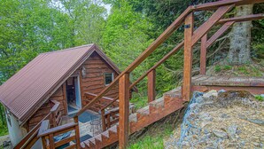A rustic cabin with a wooden staircase leading to a door, surrounded by lush greenery.