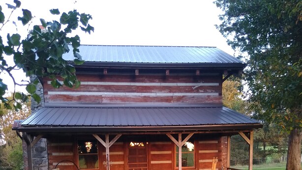 Tunnelmallinen hirsihökkelö, jossa on kuisti, kivinen perustus ja metallikatto.