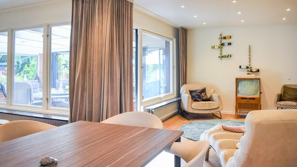 Una sala de estar con una mesa de madera, dos sillas, un sofá, un televisor y un ventanal con cortinas.