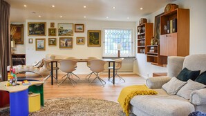 Una sala de estar con sofá, una mesa de centro y una zona de comedor con sillas y una mesa. Hay cuadros enmarcados en la pared y una estantería con vasos.