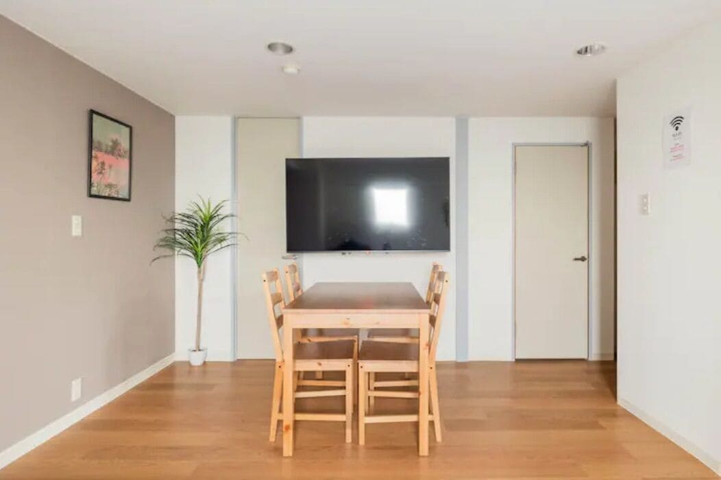 A dining area with a wooden table and chairs, a potted plant, a flat-screen TV mounted on the wall, and a door to the right.